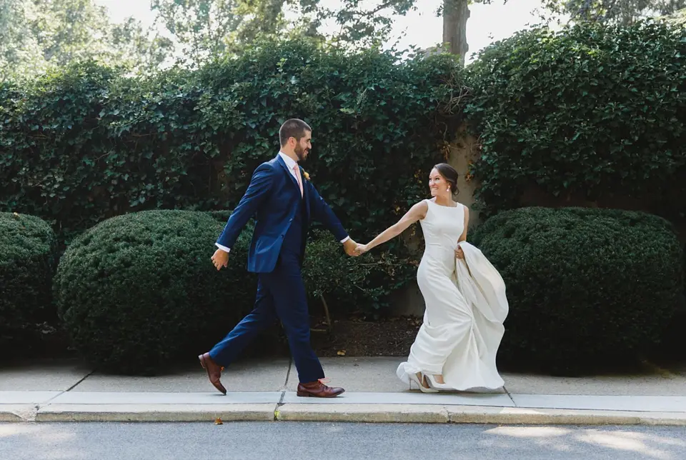 Groom in a navy suit holding the bride's hand as they walk along a sidewalk past green hedges
