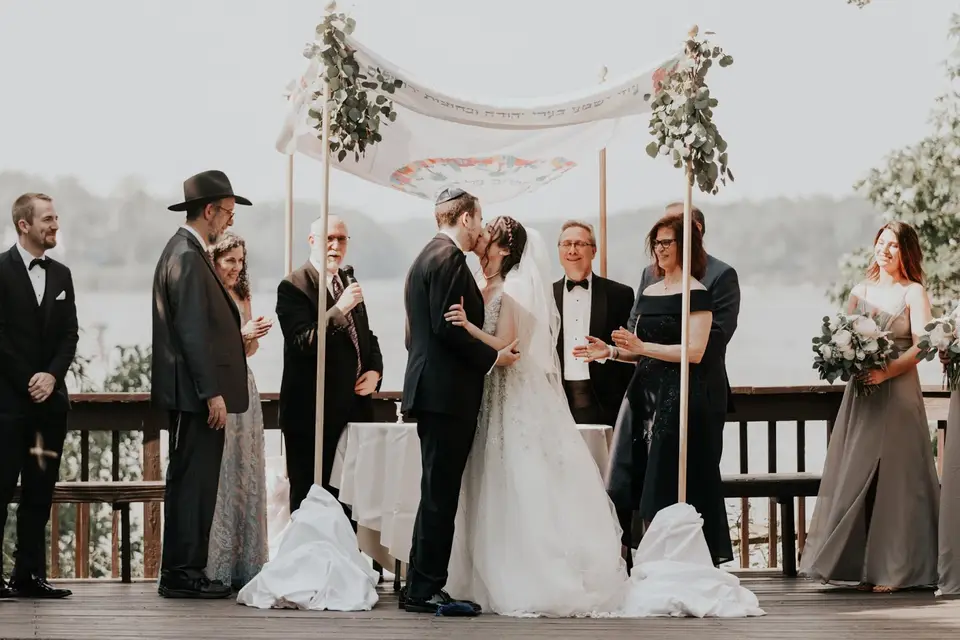 Zephan and Mollye kissing under a floral chuppah on a lakeside deck with guests and officiants around