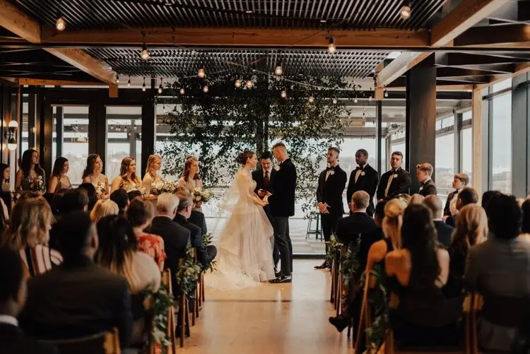 Couple exchanging vows at an indoor ceremony with guests seated and wedding party standing under wooden beams