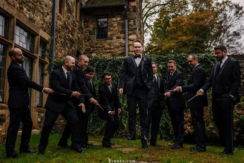 Groomsmen in dark suits pointing wands at each other on a lawn in front of a stone building