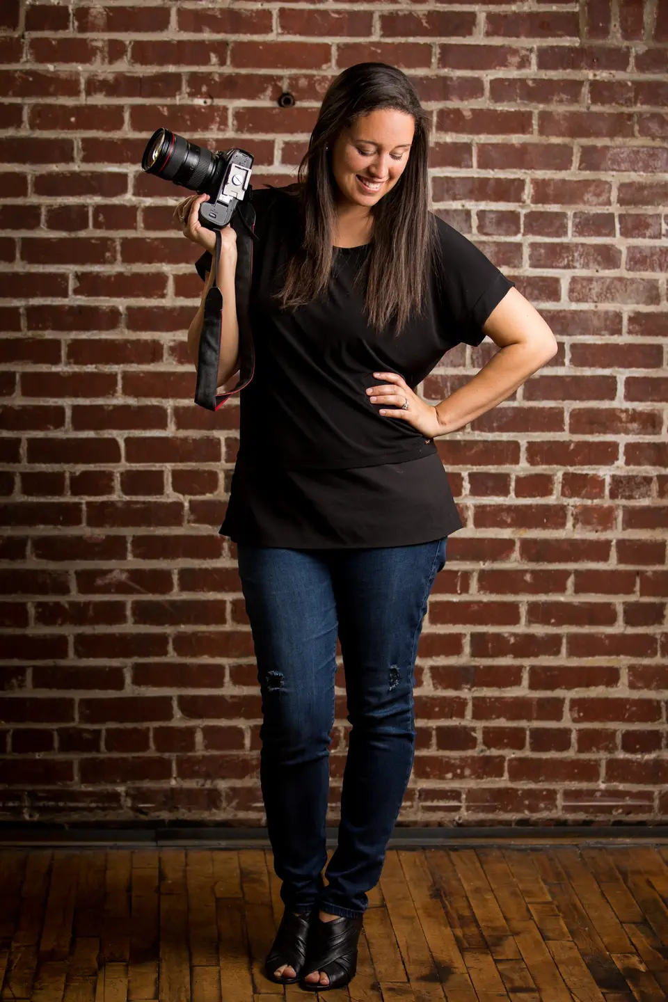 Woman photographer in a black t-shirt and jeans holding a camera, standing against a red brick wall