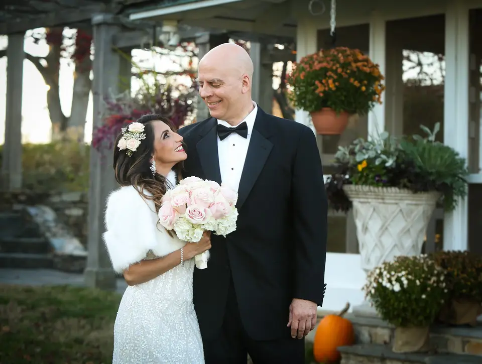 Bride in a white fur wrap and groom in a black tuxedo smile outdoors near a stone patio