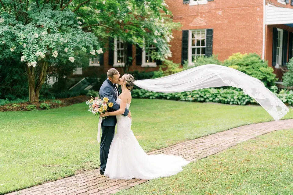 Bride and groom kissing on a brick path as her long lace veil blows out behind her, with a brick house behind