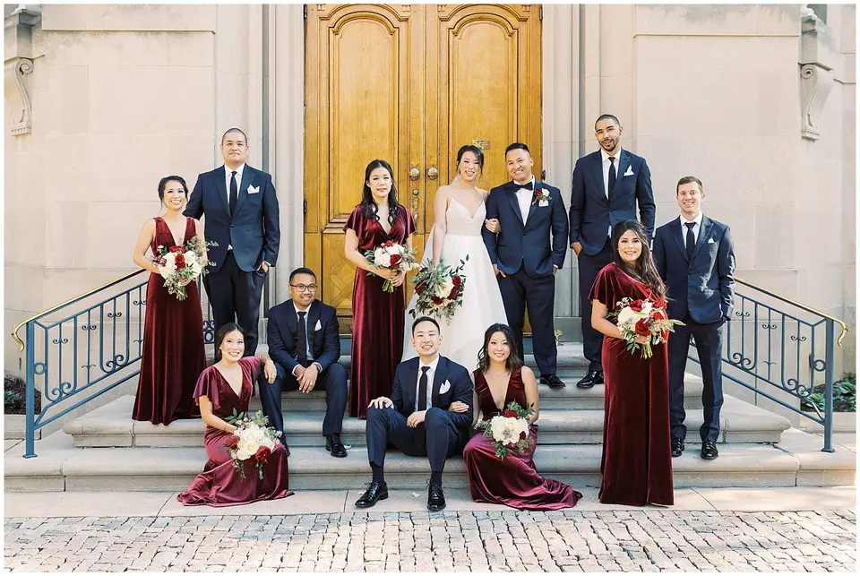 Bride and groom with bridesmaids in burgundy velvet dresses and groomsmen posed on steps by wooden doors