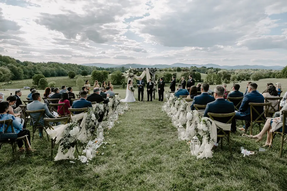 Wedding ceremony on a grassy hilltop with guests seated along flower-lined aisles and mountains in the distance