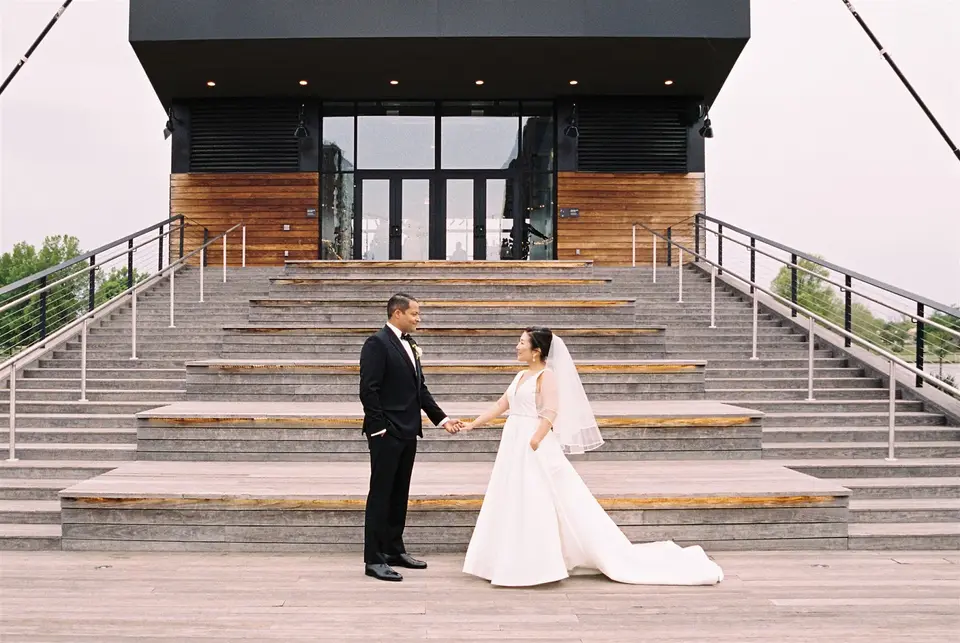 Bride and groom holding hands on wide wooden steps in front of a modern waterfront building