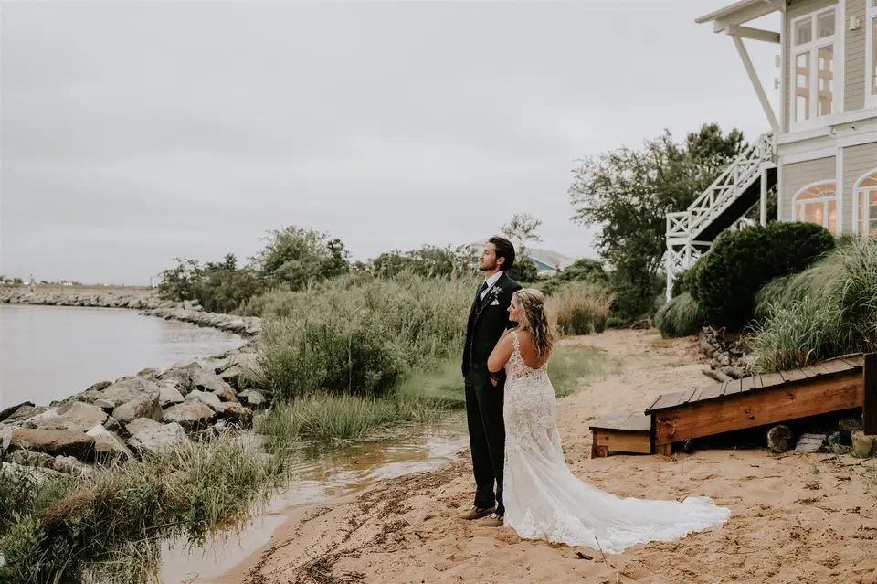 Bride and groom stand on a sandy beach by a rock-lined shore with a waterfront house behind them