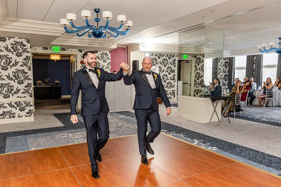 Two grooms in black tuxedos holding hands as they enter a ballroom with a wooden dance floor