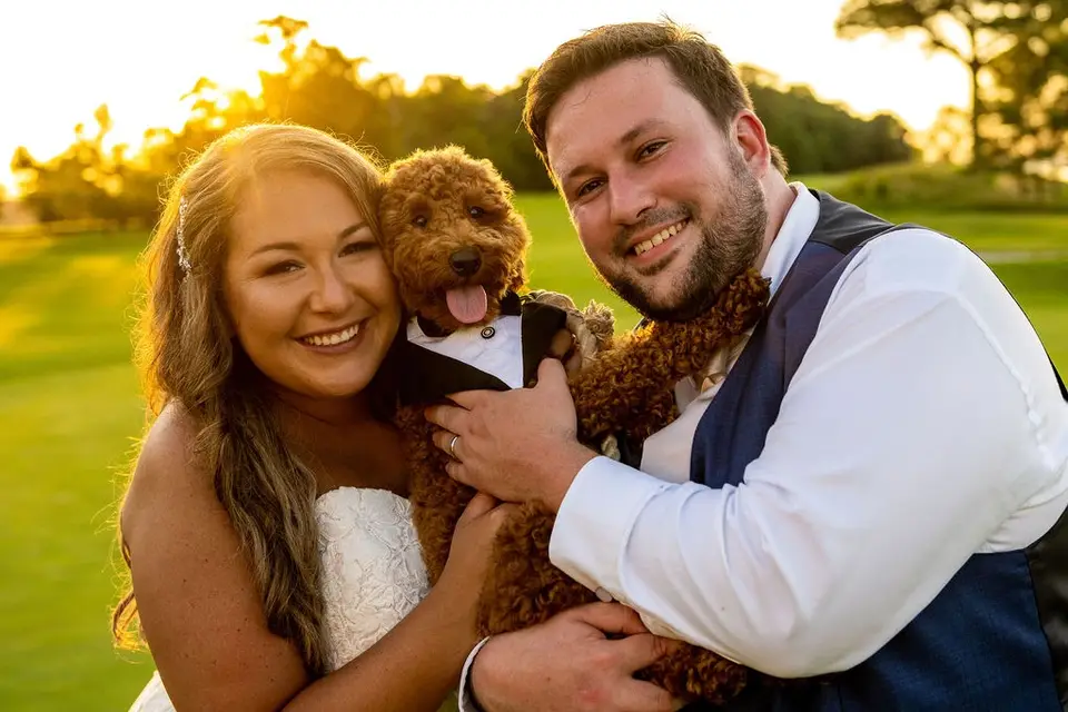 Bride and groom smiling and holding their small curly-haired dog in a tuxedo collar at golden hour