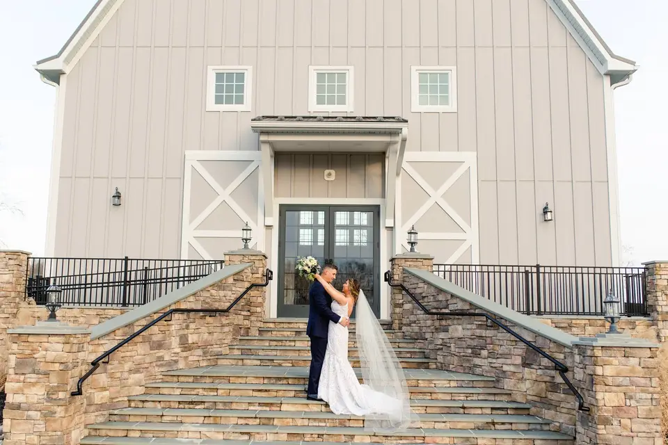 Couple embracing on stone steps in front of a large gray barn venue with a long veil