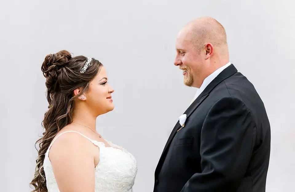 Bride and groom smiling at each other face to face against a plain gray background
