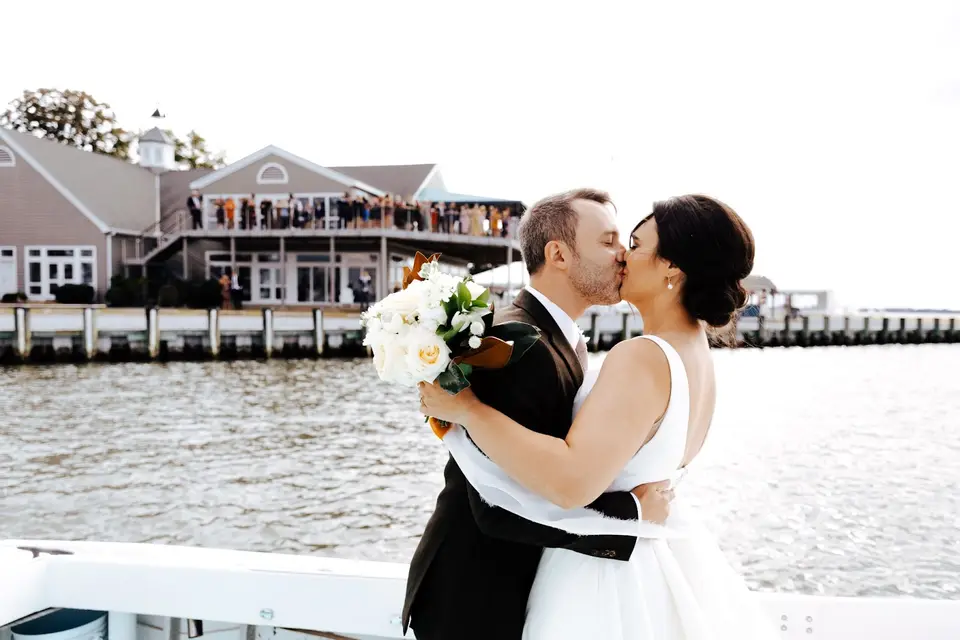 Bride and groom kissing on a boat deck with a waterfront building and wedding guests on a deck behind them