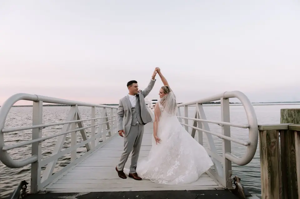 Bride in a lace gown and groom in a gray suit dancing on a wooden pier at sunset over the water