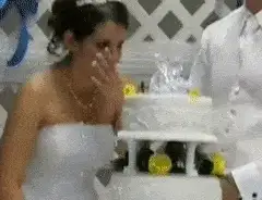 Crying bride in a strapless gown wiping her eyes in front of a damaged wedding cake