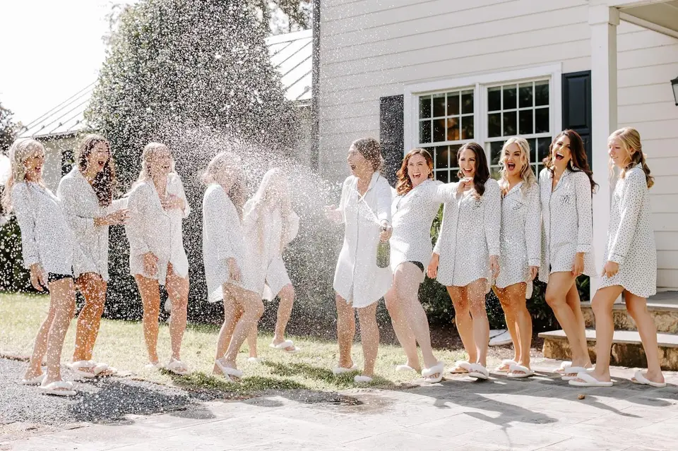 Bridesmaids in matching white shirts laughing as champagne sprays across a lawn