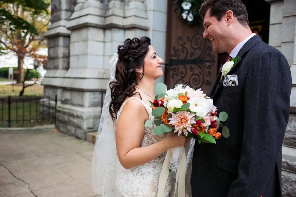Bride in a veil and groom smiling at each other outside a stone church with a floral bouquet in the foreground