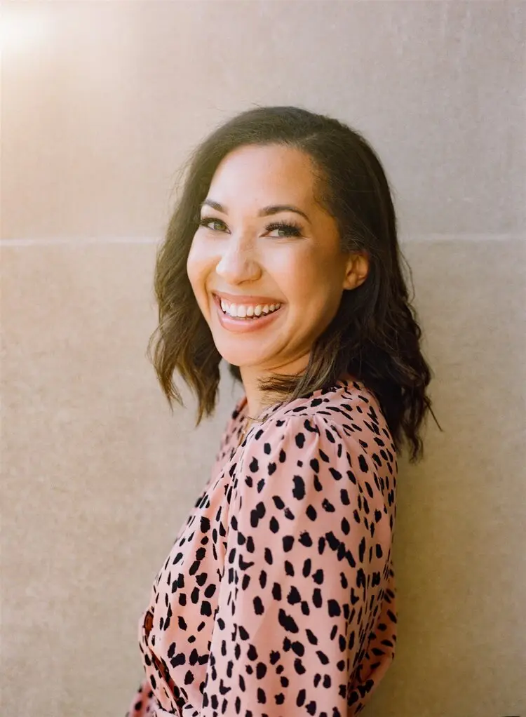 Smiling woman with dark wavy hair in a pink leopard-print top standing against a stone wall