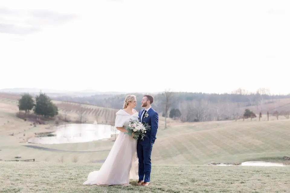 Bride in a white wrap and groom in a navy suit stand on a vineyard hillside with a pond behind