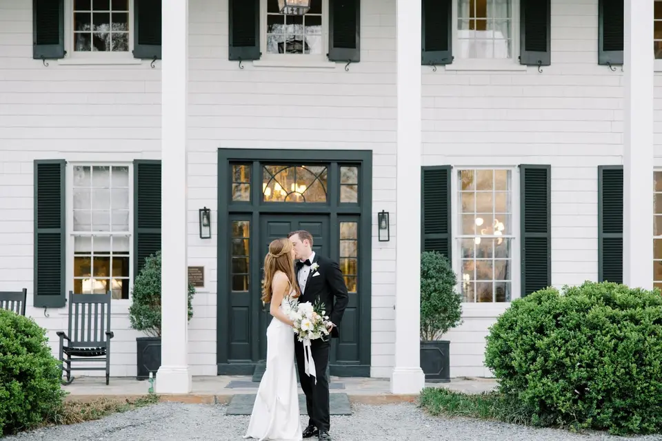 Bride and groom kissing on the front step of a white house with dark green doors and shutters