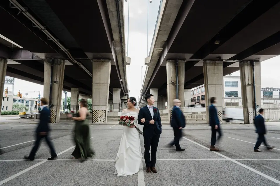 Bride and groom stand together under a highway overpass while blurred guests walk past them
