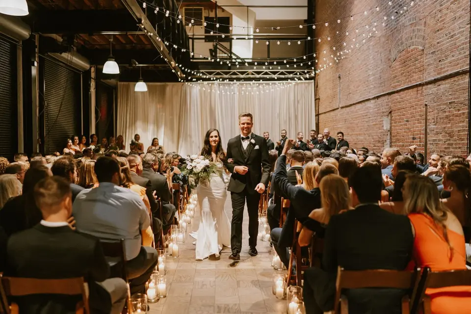 Bride and groom walking up a candlelit aisle between seated guests in a brick industrial venue