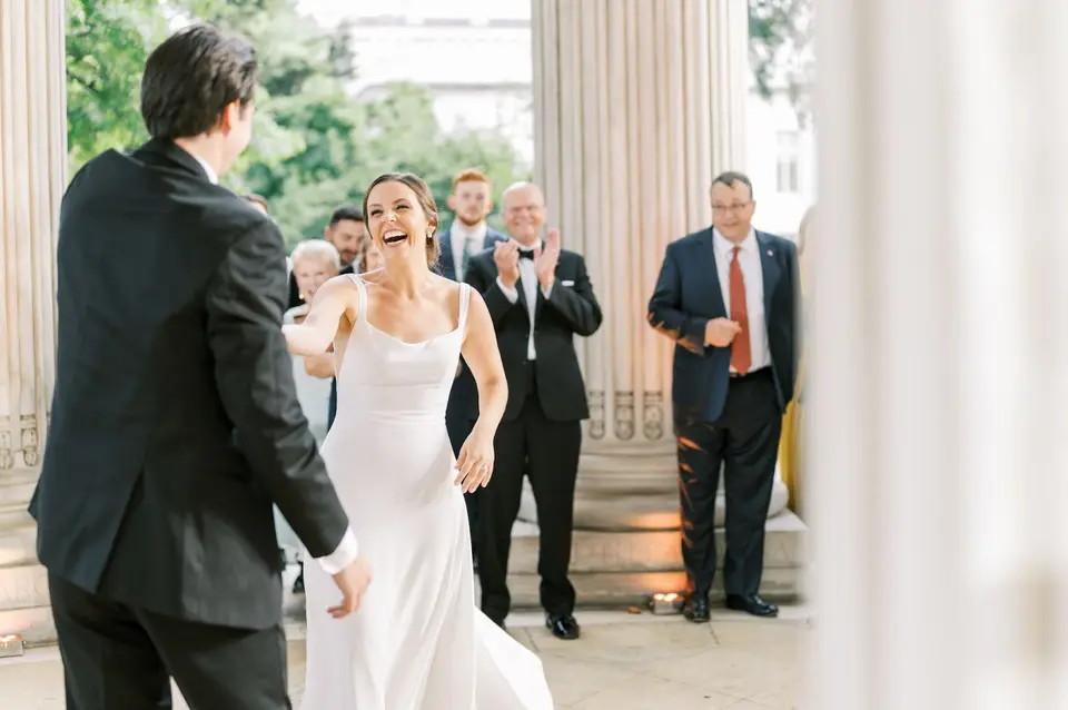 Bride dancing with her arms out and smiling toward the groom while guests watch on stone steps