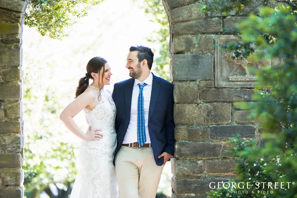 Couple smiling at each other while leaning against a stone archway in a garden