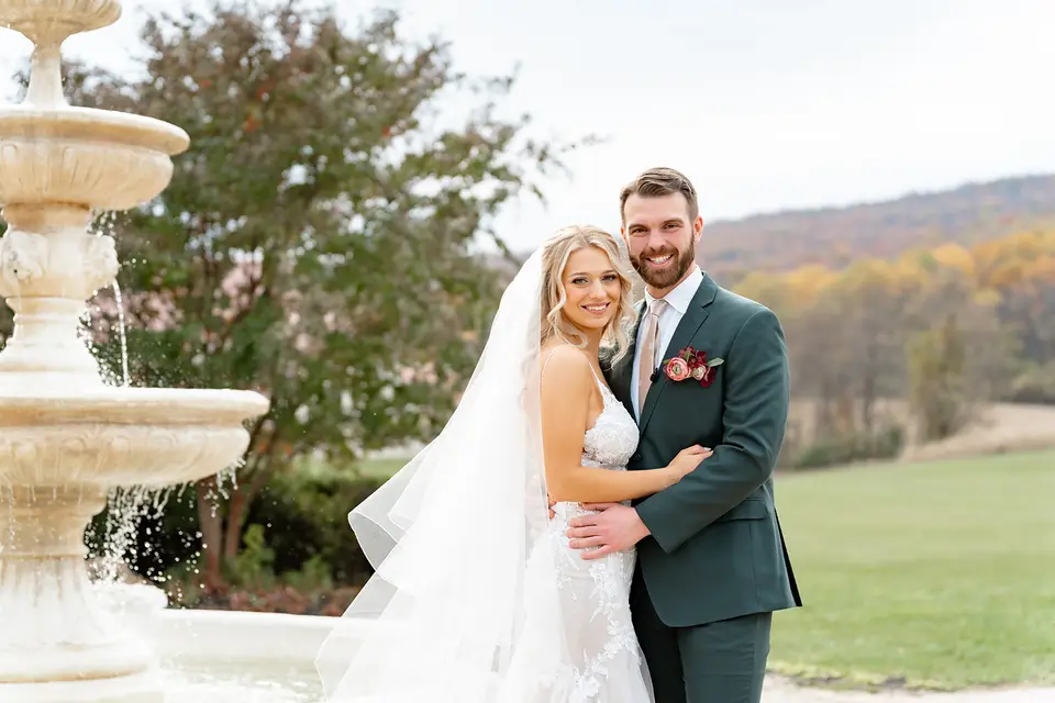 Bride and groom embracing beside a stone fountain with autumn foliage and hills behind them