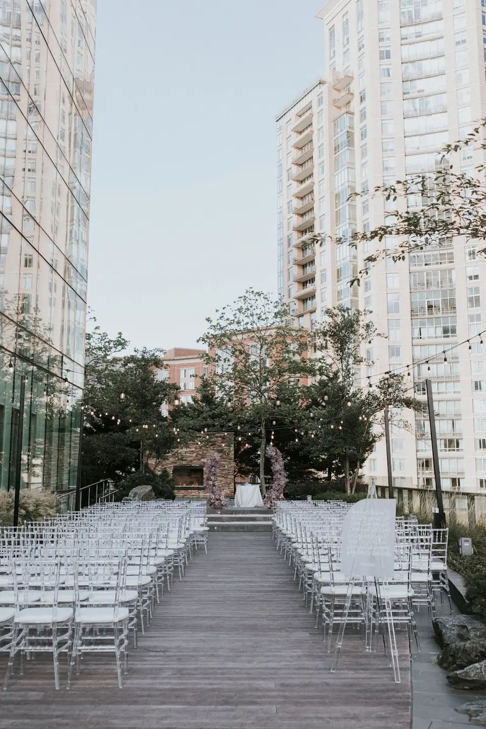 Rows of clear chairs on a wooden rooftop deck set for a ceremony between city high-rises
