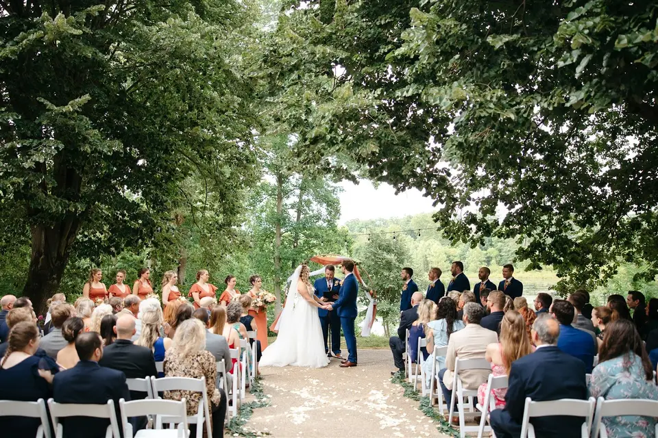 Outdoor wedding ceremony under a wooden arch in a wooded setting, couple at the front and guests seated in rows