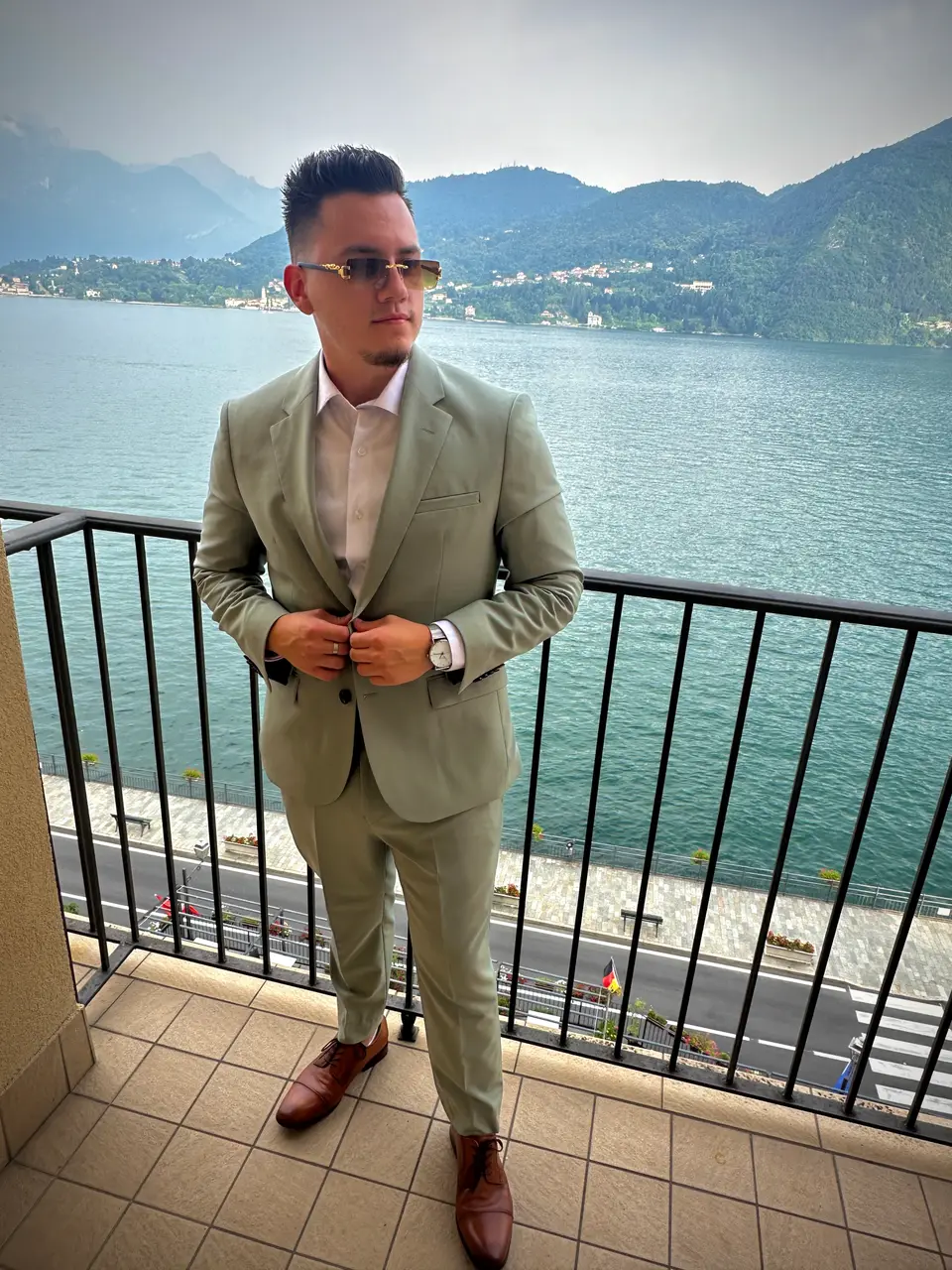 Man in a sage green suit and sunglasses standing on a balcony overlooking a lake and mountains
