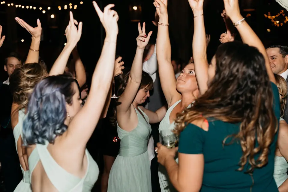 Bridesmaids in sage green dresses dancing with arms raised at a wedding reception