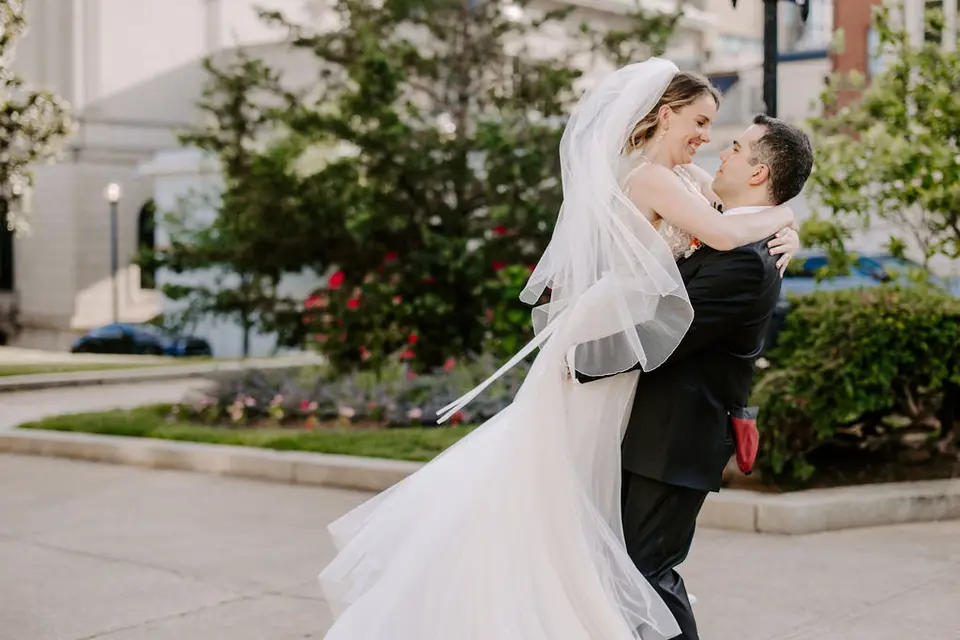 Bride and groom embracing as he lifts her, her veil flowing, on a sidewalk after their Peabody Library wedding