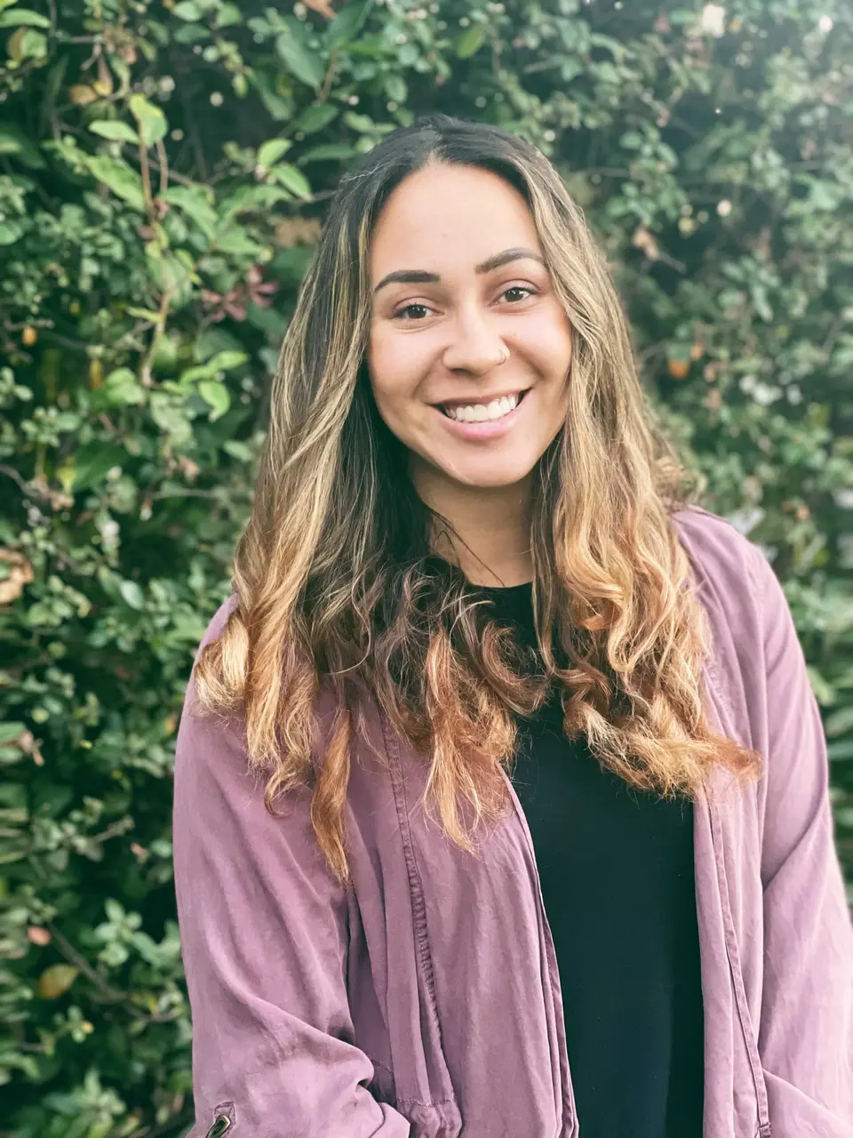 Smiling woman with long wavy hair in a lavender jacket standing in front of green foliage